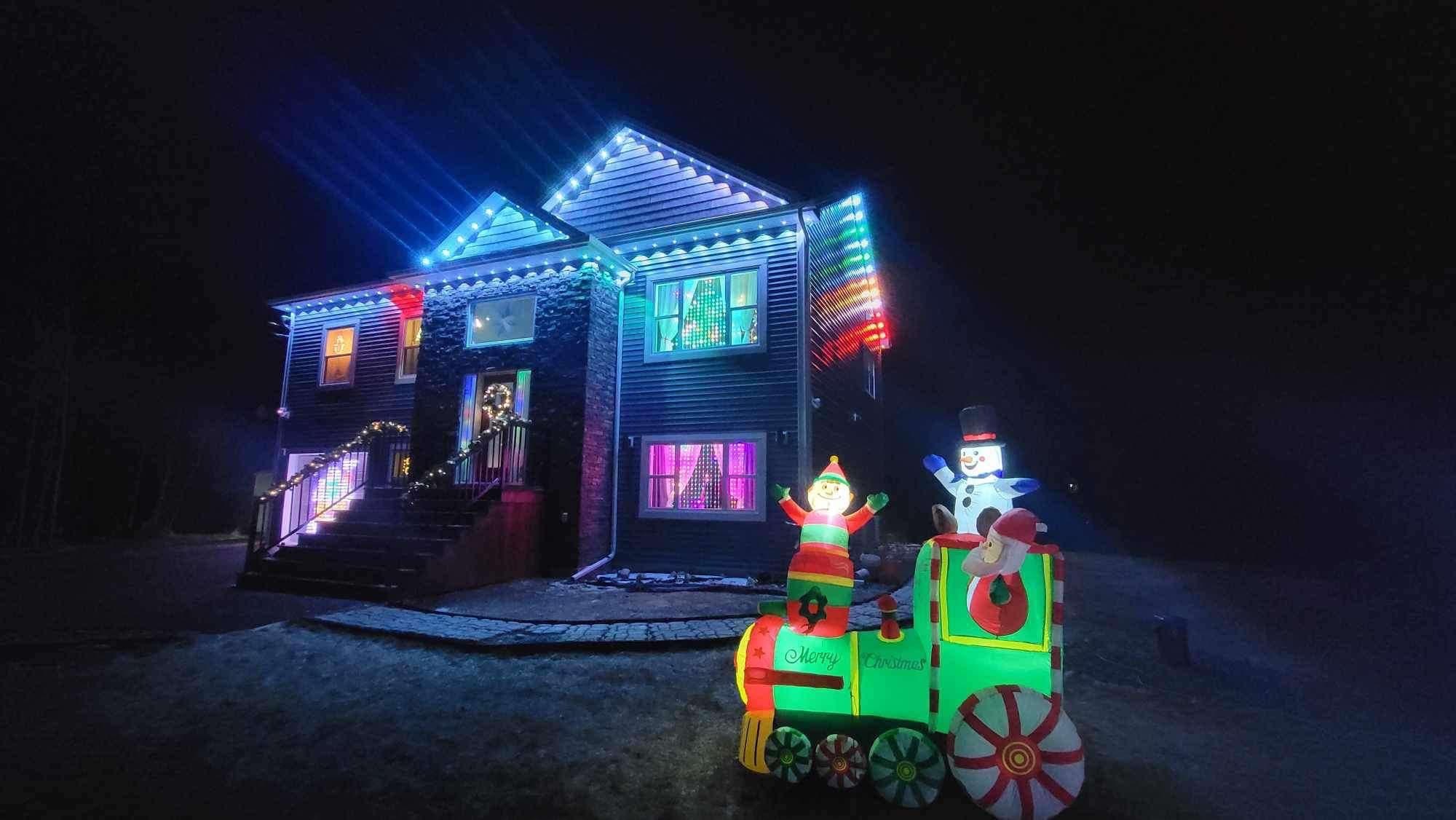 Two-storey house lit in multicolour roofline lights at Christmas, with an inflatable train on the lawn